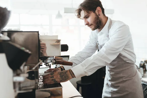 Young man working with coffee machine at the bar counter in coffee shop. Barista in apron and white shirt making coffee by coffee machine in restaurant. Boy working as barista at cafe — Stock Photo, Image