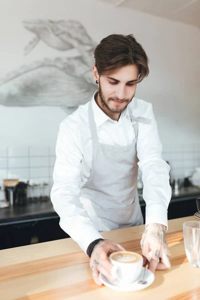 Retrato de un joven en delantal dando una taza de café mientras está de pie en el mostrador del bar en la cafetería. Barista haciendo una taza de capuchino fresco en la cafetería. Niño trabajando como barista en restaurante — Foto de Stock