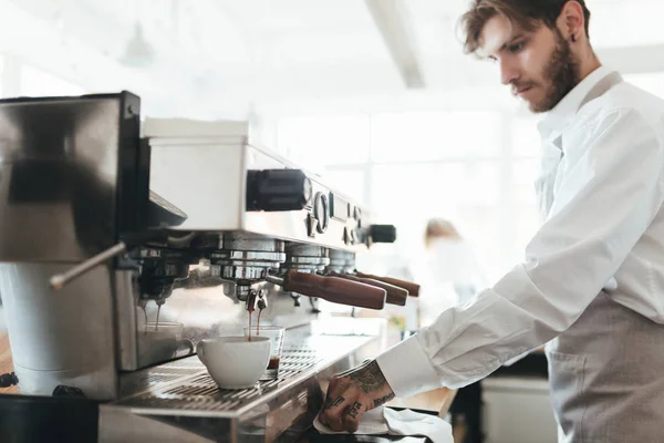 Un uomo premuroso che lavora con la macchina del caffè in caffetteria. Giovane barista in grembiule e camicia bianca che fa caffè da macchina da caffè al bancone del ristorante. Ragazzo che lavora come barista al bar — Foto Stock