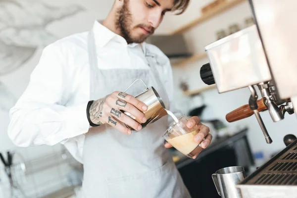 Retrato de un joven camarero haciendo una taza de café fresco en el mostrador del bar en la cafetería. Chico fresco en delantal y camisa blanca trabajando como barista en restaurante . — Foto de Stock