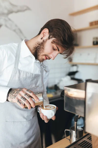 Portrait of young man working as barista and making coffee in restaurant. Barista in apron and white shirt standing with cup of coffee at his workplace in coffee shop — Stock Photo, Image