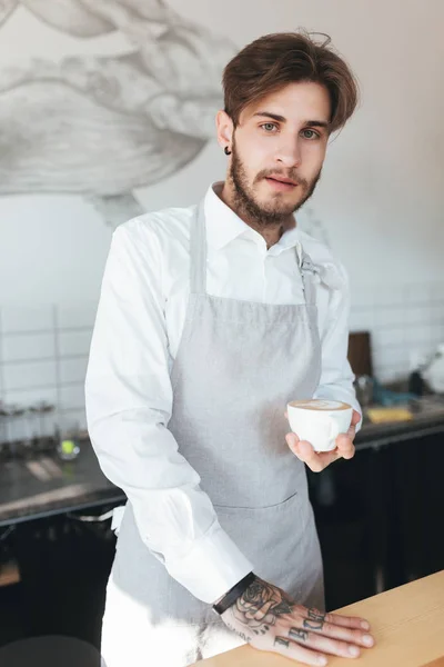 Ritratto di giovane barista che guarda in camera con una tazza di caffè in mano nel ristorante. Uomo in grembiule e camicia bianca in piedi con una tazza di caffè sul posto di lavoro in caffetteria — Foto Stock