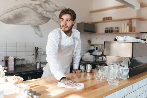 Young man thoughtfully looking aside and wiping counter at his workplace in restaurant. Portrait of barista in apron and white shirt standing at the counter in coffee shop — Stock Photo, Image