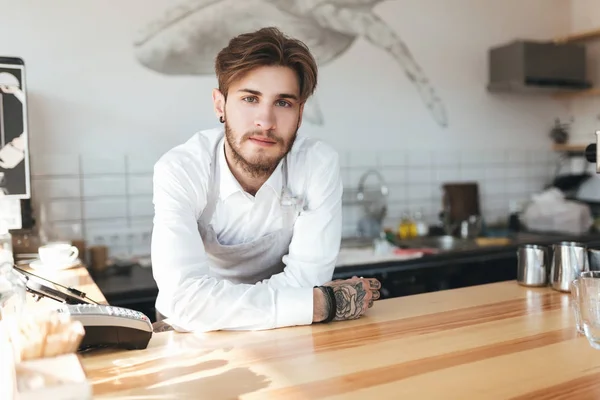 Retrato de un joven camarero parado en el mostrador del restaurante. Hombre con delantal y camisa blanca apoyando sus manos en el mostrador mirando en la cámara en el trabajo en la cafetería — Foto de Stock