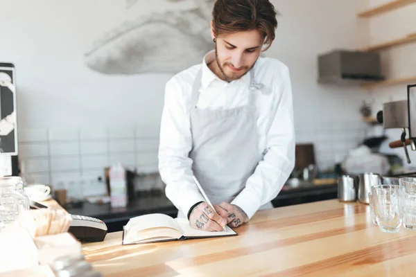 Young barista in apron and white shirt writing in notebook on workplace at counter in restaurant. Barista thoughtfully making notes at work in coffee shop. Portrait of young barista on workplace — Stock Photo, Image