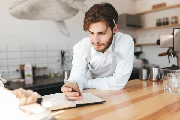 Portrait of young barista leaning his hands on counter happily looking in his mobile phone in hand at cafe. Boy in apron and white shirt with pencil behind ear standing at the counter in coffee shop — Stock Photo, Image