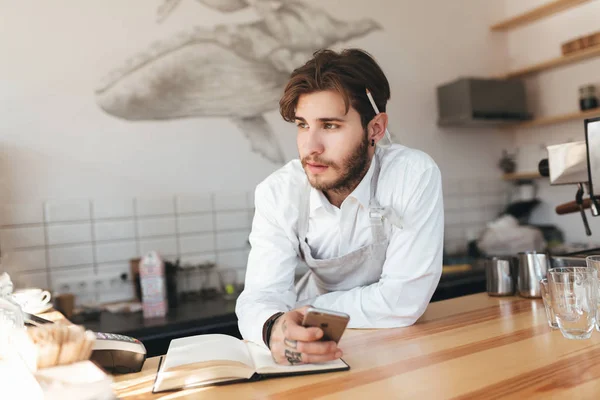 Portrait of young barista leaning his hands on counter thoughtfully looking aside with mobile phone in hand at cafe. Man in apron and white shirt standing at the counter in coffee shop — Stock Photo, Image