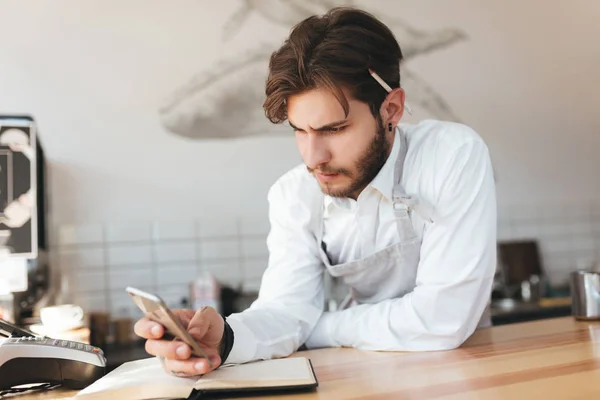 Portrait of young barista leaning his hands on counter thoughtfully looking in his mobile phone in hand at cafe. Man in apron and white shirt standing at the counter in coffee shop — Stock Photo, Image