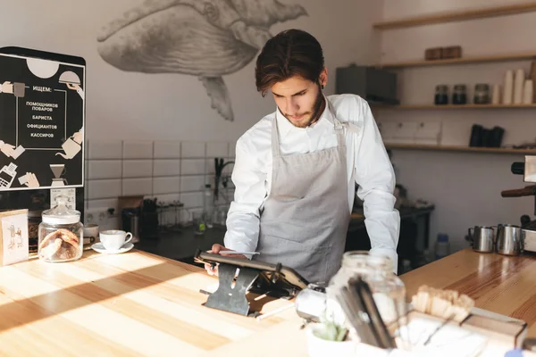 Young barista in apron and white shirt using cash counter in coffee shop. Thoughtful man working with terminal at the counter on his workplace in restaurant — Stock Photo, Image