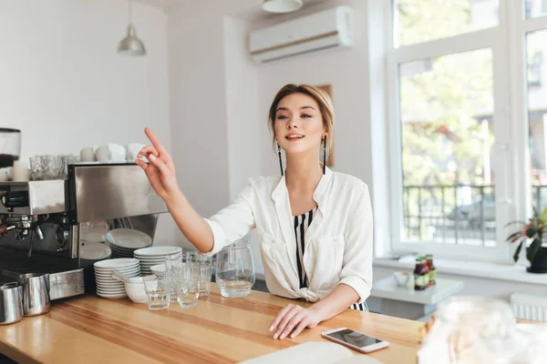 Young beautiful girl sitting at the counter in coffee shop and calling barista. Portrait of smiling lady with blond hair happily looking aside ready to make order in restaurant — Stock Photo, Image