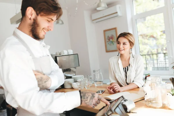 Jonge barista in schort en wit hemd met behulp van cash counter in de coffeeshop. Mooi lachende meisje met blond haar zitten aan de balie in restaurant. Barista en mooi meisje gelukkig praten op café — Stockfoto