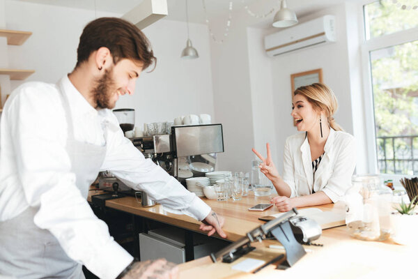 Young barista in apron and white shirt working at the counter in coffee shop. Nice smiling girl sitting at the counter at cafe and showing peace gesture. Barista and girl happily talking at cafe