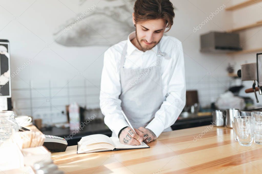 Young barista in apron and white shirt writing in notebook on workplace at counter in restaurant. Barista thoughtfully making notes at work in coffee shop. Portrait of young barista on workplace