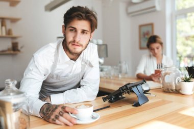 Önlük ve Beyaz gömlekli düşünceli barista portresi işyeri kafede kahve fincan ile. Sersem bir kenara güzel kız ise sayaç cep telefonu kullanarak özgeçmişlerine bakarak Barista 