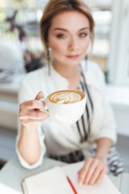 Restoran ile defter masada oturan ve kahve içme sarı saçlı genç güzel kız portresi. Kadın el fincan cappuccino kahve dükkanında tutarak yukarı kapatın 