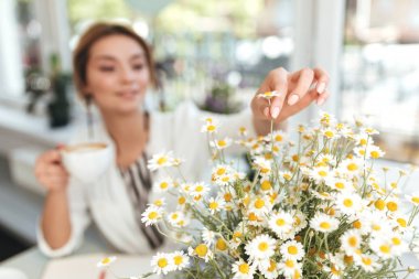 Elinde fincan cappuccino kahve dükkanında oturmuş sarı saçları ve küçük chamomiles büyük buket kızla. Kadın eli papatya çiçeği buket dokunmadan kapatın