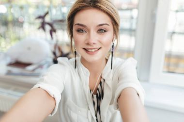 Güzel genç kız onu ön kamera içinde hareket eden telefon fotoğraf yapma. Çok gülümseyen kadın mutlu bir şekilde kamera seyir restoranda kulaklık sarışın saçlı. Serin kız giyiyor beyaz gömlek
