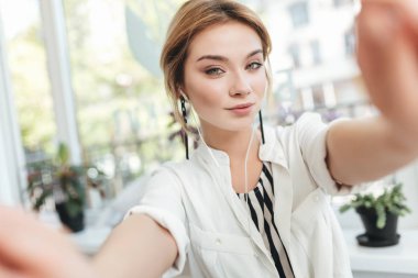 Güzel genç kız onu ön kamera içinde hareket eden telefon fotoğraf yapma. Düşünceli kamera seyir restoranda kulaklık sarışın saçlı hoş bir bayan. Serin kız giyiyor beyaz gömlek