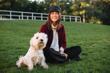 Siyah kap ve oldukça küçük köpeği ile parkta çim üzerinde oturan ve mutlu bir şekilde fotoğraf makinesindeki izole seyir ceket genç güzel Bayan portresi