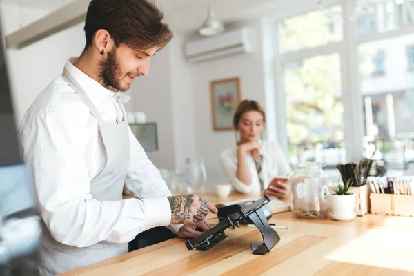Νέοι χαμογελώντας barista στην ποδιά και το λευκό πουκάμισο χρησιμοποιώντας μετρητά μετρητή στο κατάστημα καφέ ενώ στοχαστικό κορίτσι σε φόντο που κάθεται στον πάγκο με το κινητό τηλέφωνο. Δροσερό αγόρι εργάζεται ως barista στο καφενείο — Φωτογραφία Αρχείου