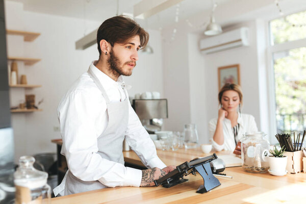 Young pensive barista in apron and white shirt using cash counter in restaurant while thoughtful girl on background sitting at the counter with mobile phone. Cool boy working as barista in coffee shop