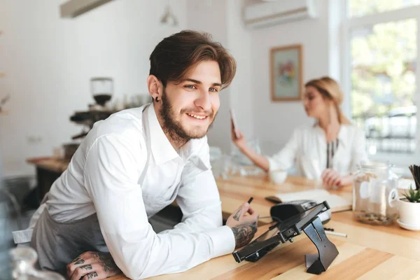 Portrait of smiling barista in apron and white shirt on workplace in coffee shop. Barista happily looking aside standing at the counter while pretty girl on background using her mobile phone at cafe — Stock Photo, Image