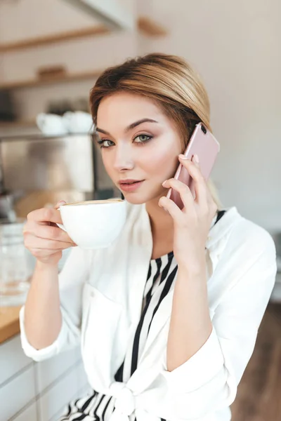 Портрет милої дівчини, яка дивиться в камеру, сидячи за прилавком і п'ючи каву з мобільним телефоном в руках у кав'ярні. Красива дама з світлим волоссям розмовляє на мобільному телефоні в кафе — стокове фото