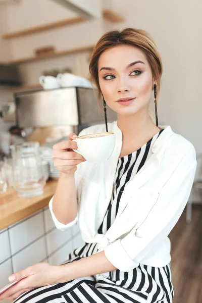 Portret dziewczyny piękne, starannie patrząc na bok, siedząc przy kasie i picia kawy w kawiarni. Ładna Pani, blond włosy, w białej koszuli siedzi w restauracji z kubkiem w ręku — Zdjęcie stockowe