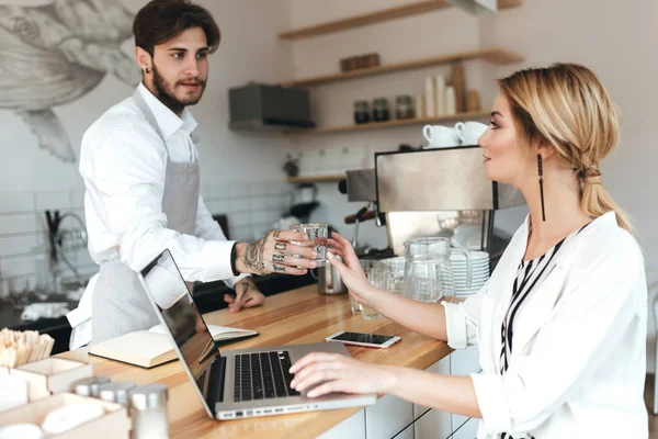 Jovem barista de avental e camisa branca dando copo de água para menina bonita. Linda senhora com cabelo loiro sentado no balcão e trabalhando em seu laptop no café — Fotografia de Stock