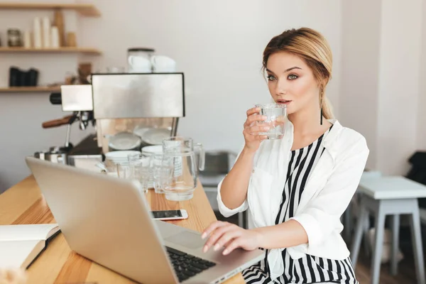 Ωραίο κορίτσι που κάθεται στον πάγκο με ποτήρι νερό στο χέρι και περίσκεψη ψάχνει στην άκρη στο εστιατόριο. Πορτρέτο του όμορφη κοπέλα με τα ξανθά μαλλιά που εργάζονται για το laptop της σε καφετέρια — Φωτογραφία Αρχείου