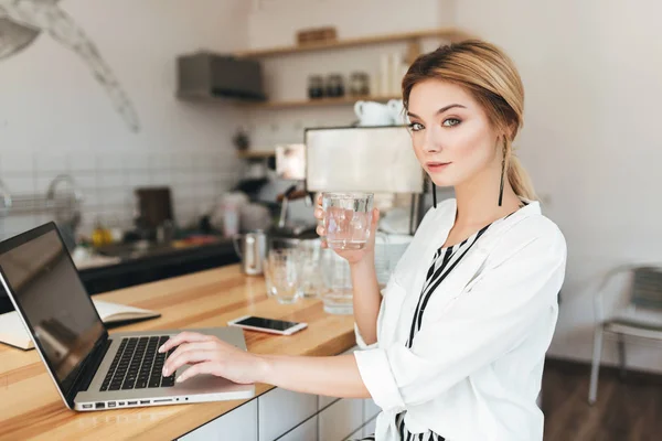 Hermosa chica sentada en el mostrador con un vaso de agua en la mano y mirando cuidadosamente en la cámara en el café. Bella dama con el pelo rubio trabajando en su portátil en la cafetería —  Fotos de Stock