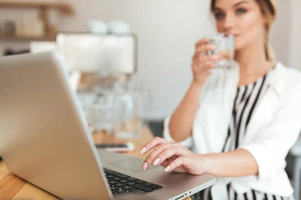 Una chica guapa con el pelo rubio bebiendo agua en el restaurante. Hermosa chica con camisa blanca trabajando en su computadora portátil en la cafetería. Primer plano mano señora escribiendo en el ordenador en la cafetería —  Fotos de Stock