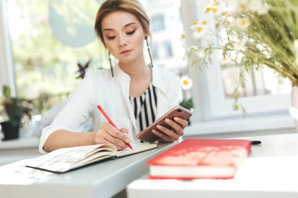 Retrato de menina bonita sentada no restaurante e escrevendo em seu caderno. Menina bonita com cabelo loiro segurando telefone celular na mão e escrevendo notas no café. Senhora agradável estudando no restaurante — Fotografia de Stock