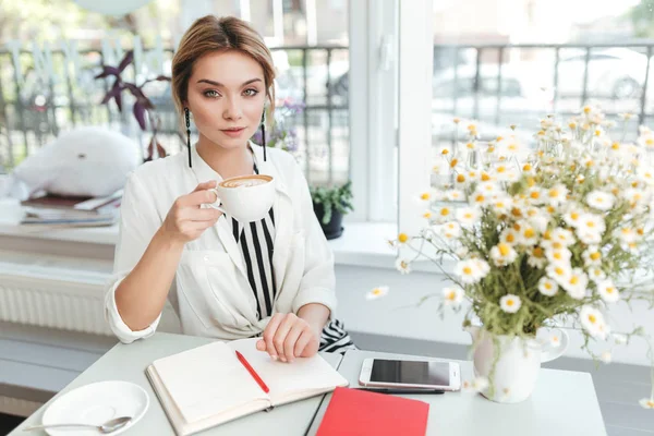 Portrait of young girl looking in camera with cup of coffee in hand at cafe. Beautiful lady with blond hair sitting in coffee shop with mobile phone,notebook and bouquet of flowers on table — Stock Photo, Image