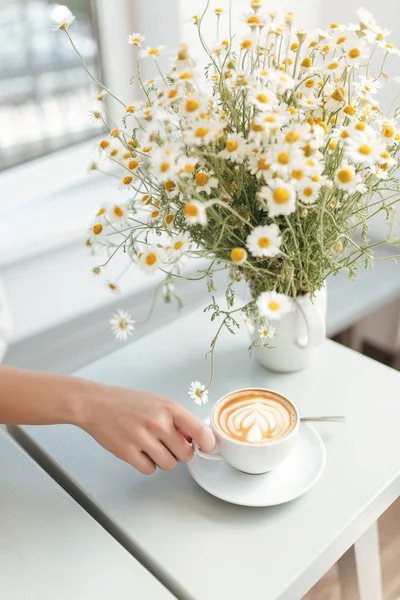 Hermosa foto de cerca de flores de manzanilla y taza blanca de capuchino en la mesa en la cafetería. Mujer mano sosteniendo la taza de café en el restaurante —  Fotos de Stock