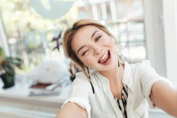 Gadis muda yang cantik membuat foto lucu di kamera depan di ponsel. Wanita cantik dengan rambut pirang di earphone dengan senang hati melihat kamera di restoran. Gadis keren mengenakan kemeja putih — Stok Foto