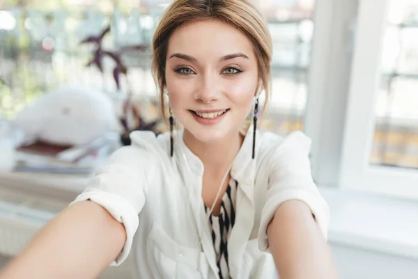 Vakker ung jente som lager bilder på frontalkameraet sitt i mobiltelefon. En smilende dame med blondt hår i øretelefoner som gledelig ser i kamera på restaurant. Kul jente i hvit skjorte – stockfoto