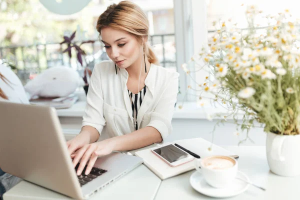 Buena chica con auriculares sentados en la cafetería y escribiendo en su computadora portátil. Retrato de una joven con el pelo rubio trabajando en el ordenador en el restaurante con teléfono móvil, portátil y taza de café en la mesa —  Fotos de Stock