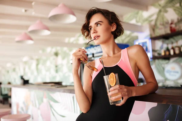 Giovane ragazza premurosa in costume da bagno alla moda appoggiata al bancone del bar sulla spiaggia con occhiali da sole e cocktail in mano. Ritratto di bella signora in piedi al bancone del bar e sognante guardando da parte — Foto Stock