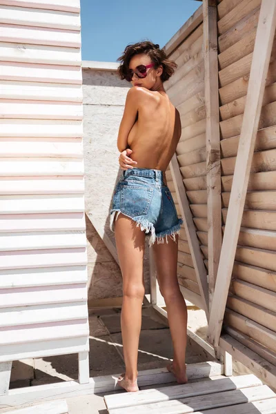 Belle fille en short denim et lunettes de soleil passant du temps sur la plage. Jeune jolie fille debout de dos tout en couvrant sa poitrine avec les mains dans les vestiaires sur la plage — Photo