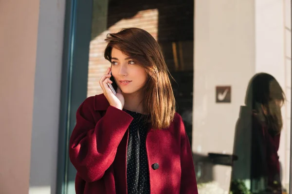 Портрет юной леди в пальто, стоящей на улице и мечтательно смотрящей в сторону, разговаривая по мобильному телефону изолированной — стоковое фото