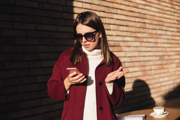 Muotokuva kaunis tyttö aurinkolasit ja takki seisoo ja huomaavaisesti etsivät hänen matkapuhelin — kuvapankkivalokuva