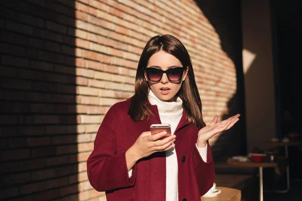 Portrett av en ung dame i solbriller og frakk mens hun sto og så på mobilen – stockfoto