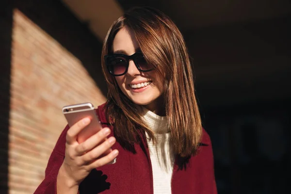 Portrett av en ung smilende dame i solbriller som står på gata og gladelig bruker mobilen isolert – stockfoto