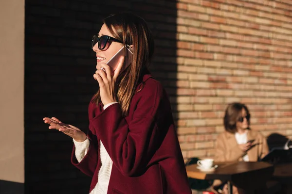 Портрет юной леди в солнечных очках и пальто, стоящей на улице, разговаривая по мобильному телефону с девушкой, что чтение книги на заднем плане — стоковое фото