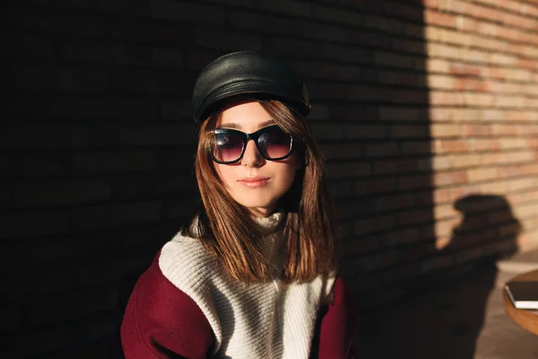 Портрет красивой девушки в солнечных очках и черной кепке, сидящей на улице и смотрящей в камеру изолированной — стоковое фото