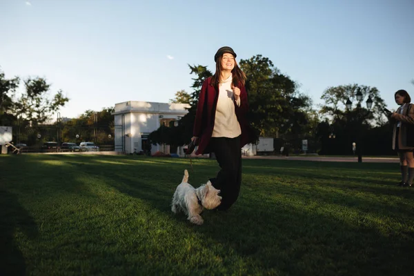 Porträtt av ung vacker dam i svart keps och kappa med glädje promenader i parken med sin lilla söta hund — Stockfoto