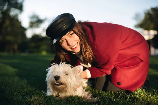 Close up foto di giovane bella signora in berretto nero e cappotto felicemente alla ricerca in macchina fotografica mentre abbraccia il suo piccolo cane carino nel parco — Foto Stock