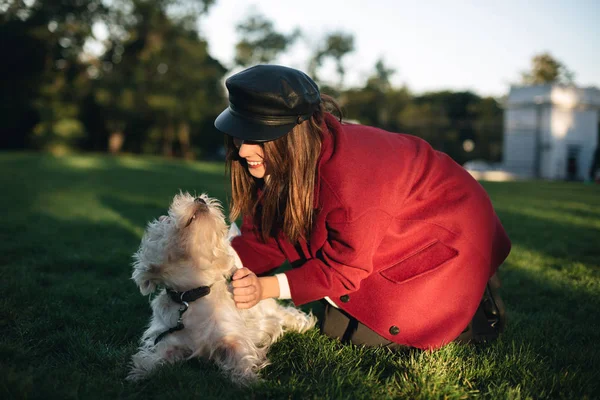 Porträtt av ung vacker dam i svart keps och coat kramas och glatt leker med hennes små söta hund i parken — Stockfoto