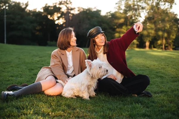 Portret dwóch pięknych dziewczyn, siedząc na trawie i biorąc selfie na telefon a spędzanie czasu z ich pies w parku — Zdjęcie stockowe